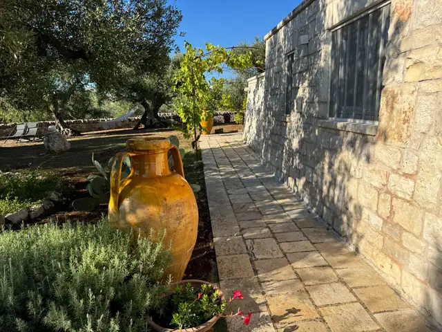 Camminamento in pietra accanto a un trullo, con anfora e piante aromatiche